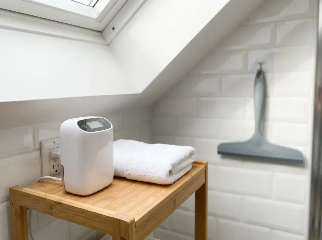 Practical small attic bathroom ideas featuring a compact dehumidifier on a bamboo shelf and a squeegee for moisture control.