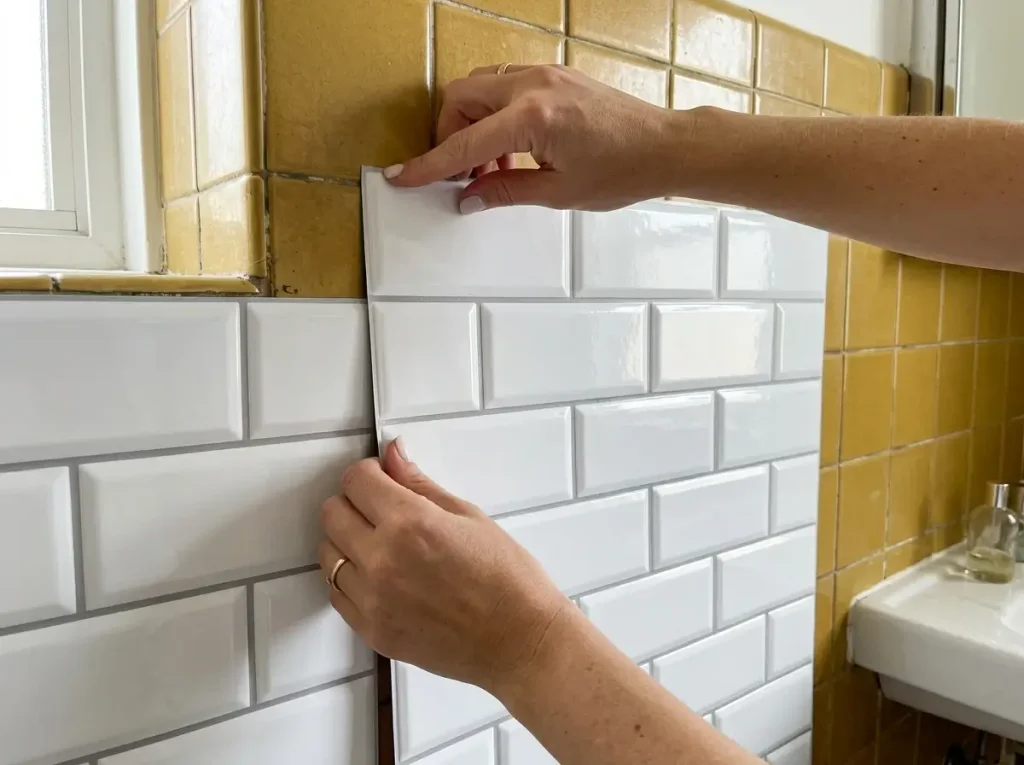 A person demonstrating how to safely cover rental bathroom tiles using 3D vinyl peel-and-stick sheets over dated yellow ceramic.
