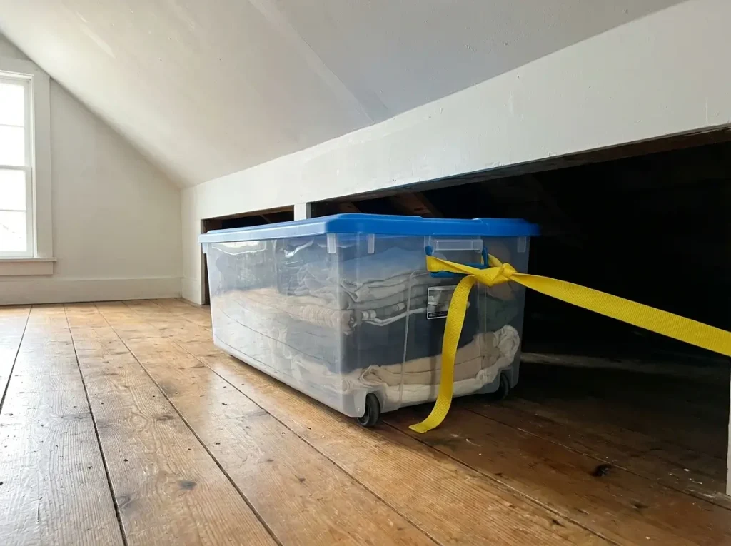 A floor-level view of practical small attic storage ideas, showing a shallow, rolling plastic bin being pulled out from underneath a sharply sloped ceiling using a yellow nylon strap.